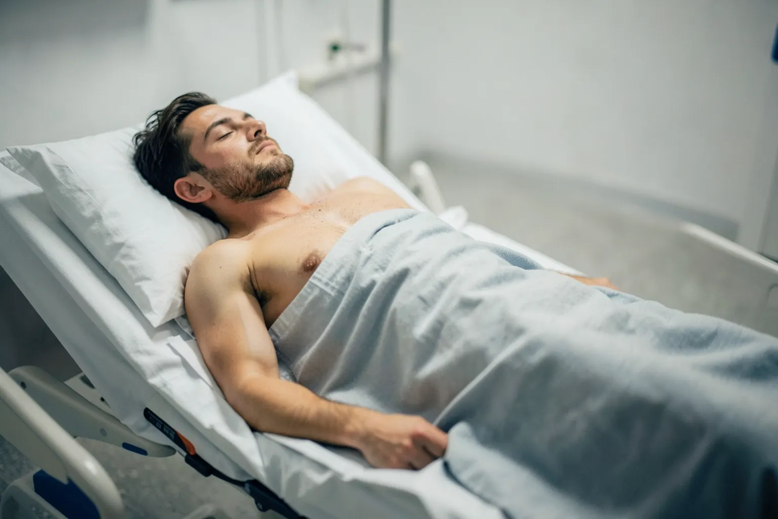 Um homem deitado em uma cama de hospital com uma expressão de dor, com um cobertor cobrindo seu corpo até a cintura.