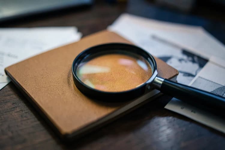 Uma lupa focando em um bloco de notas fechado com alguns papéis ao lado em uma mesa de madeira escura, plano detalhe, luz de escritório vinda de cima, sem pessoas.