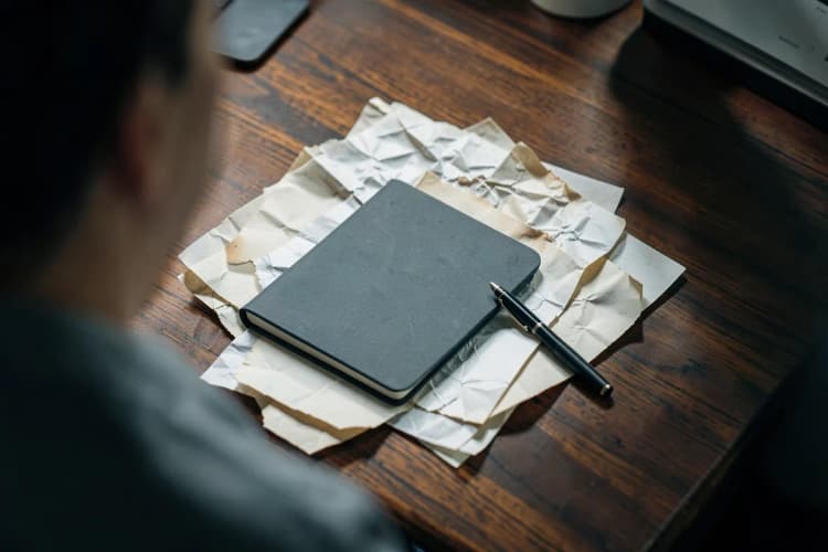 Um bloco de notas fechado e uma caneta sobre papéis amassados em uma mesa de madeira escura, plano detalhe, luz de escritório vinda de cima, sem pessoas.