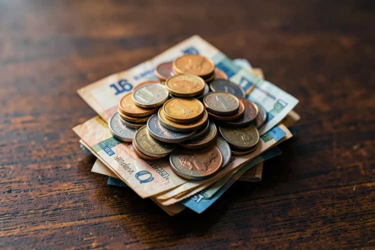 Uma pilha de moedas de diferentes valores e notas de dinheiro dobradas sobre uma mesa de madeira escura.
