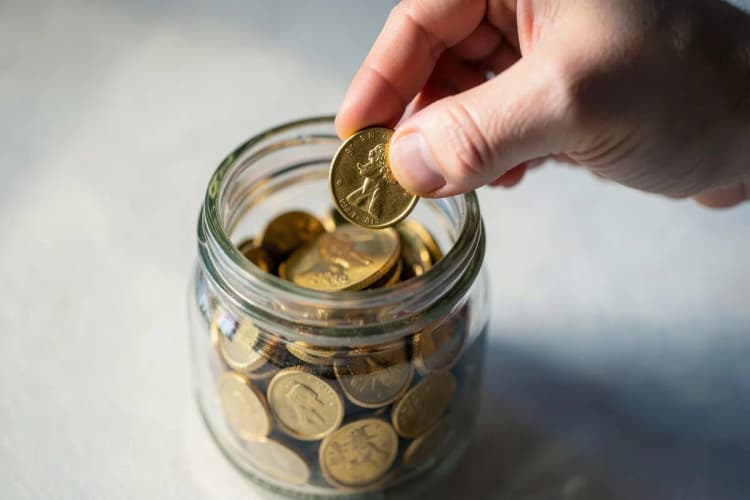 Uma mão colocando uma moeda de ouro em uma jarra de vidro transparente cheia de outras moedas.